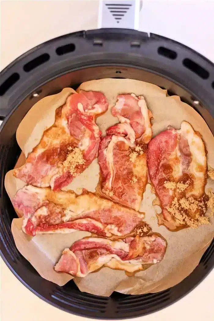 Slices of raw bacon, marbled with fat and kissed with maple syrup, are arranged on parchment paper inside an air fryer basket. The strips appear to be sprinkled with brown sugar, creating the solid base for candied bacon.
