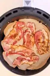 Slices of raw bacon, marbled with fat and kissed with maple syrup, are arranged on parchment paper inside an air fryer basket. The strips appear to be sprinkled with brown sugar, creating the solid base for candied bacon.