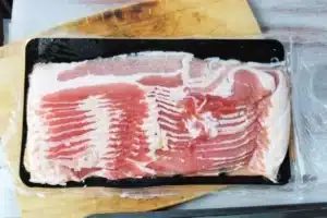 A sealed package of uncooked bacon strips is placed on a wooden cutting board.