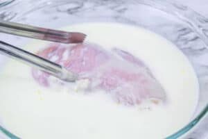 Raw chicken breast being dipped in a bowl of milk with metal tongs on a marble surface, the first step to recreating an Air Fryer McDonalds Fried Chicken Sandwich at home.