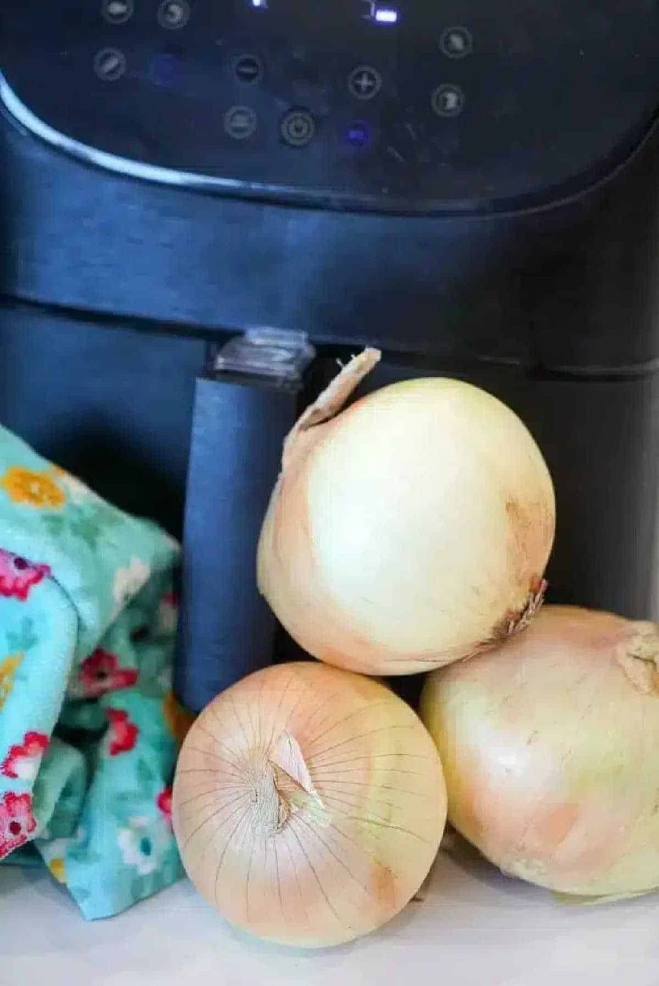 Three yellow onions sit in front of a black air fryer, solid for making crispy air fryer onions, with a colorful cloth adding a pop of color to the left.