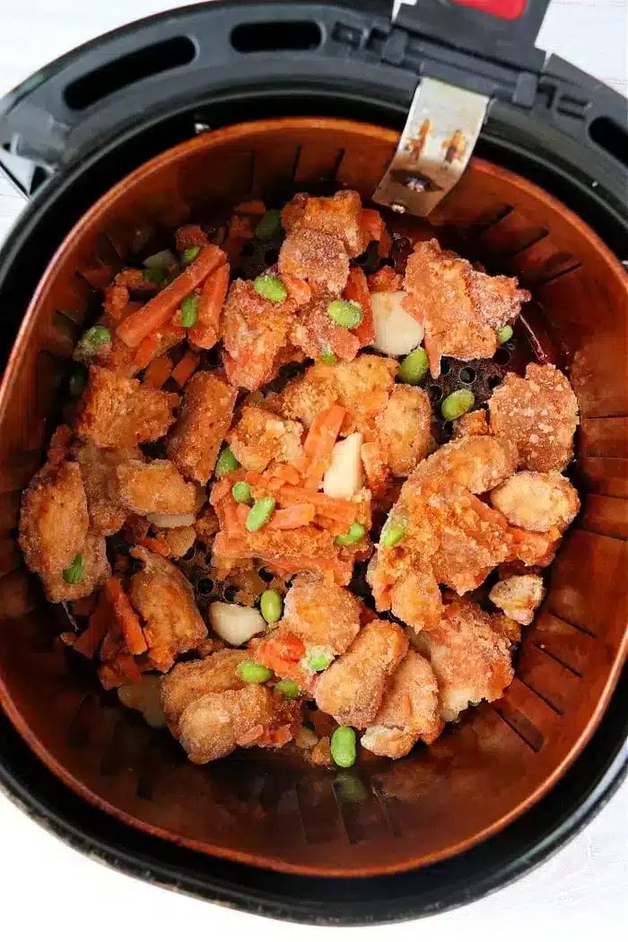 An air fryer basket filled with mixed, breaded orange chicken pieces, carrots, and green edamame beans. The food appears crispy and is evenly distributed in the basket.