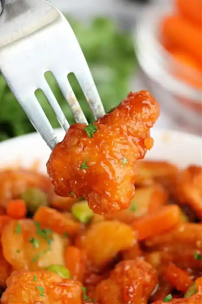 A fork holds a piece of orange-glazed chicken, perfectly crispy from the air fryer, over a bowl filled with more pieces and mixed vegetables like carrots and peas. Parsley is sprinkled on top for garnish.