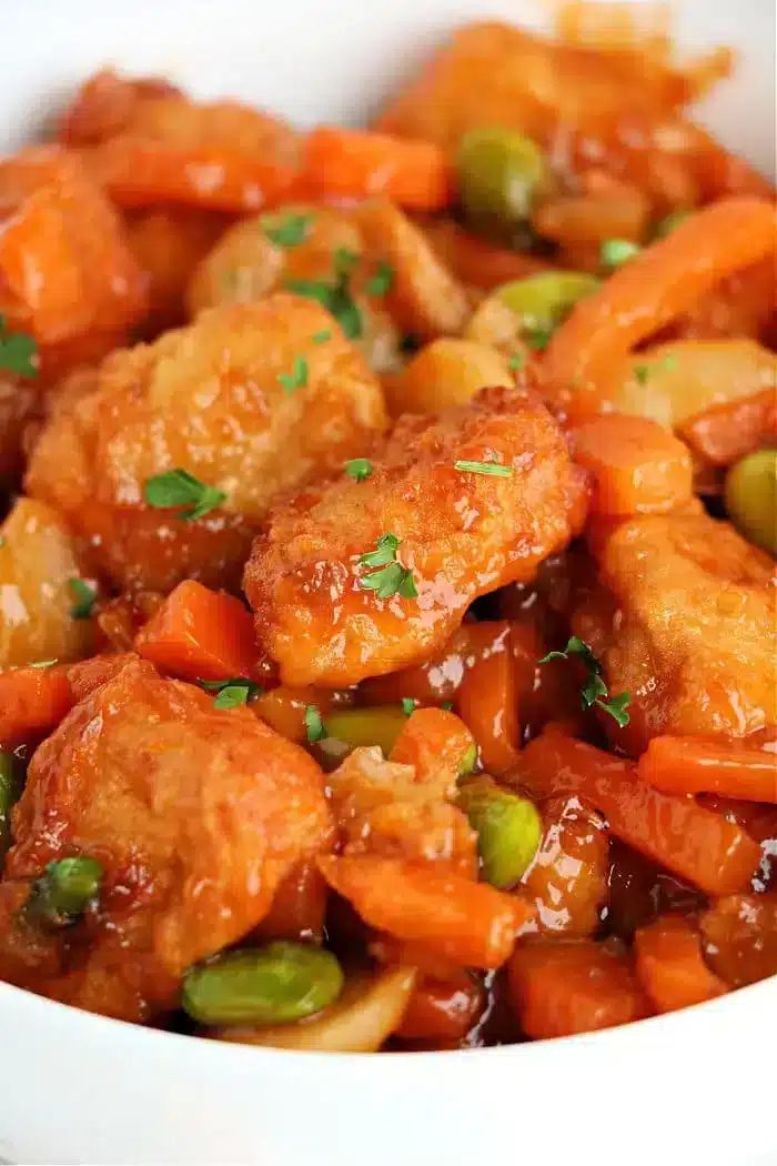 A close-up of sweet and sour air fryer chicken with crispy breaded pieces, sliced carrots, edamame, and a glossy sauce. Garnished with chopped parsley.