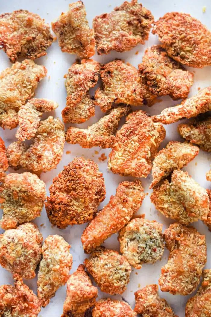 A collection of crispy, golden-brown popcorn chicken pieces arranged on a white surface. The air fryer ensures each piece achieves that textured, crunchy coating, with the pieces varying in size and shape.