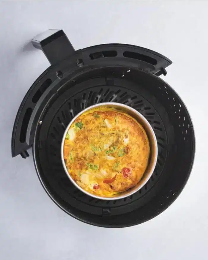 An air fryer with a removable basket contains a good quiche, featuring visible ingredients like tomatoes, green herbs, and cheese. The appliance is placed on a light-colored surface.