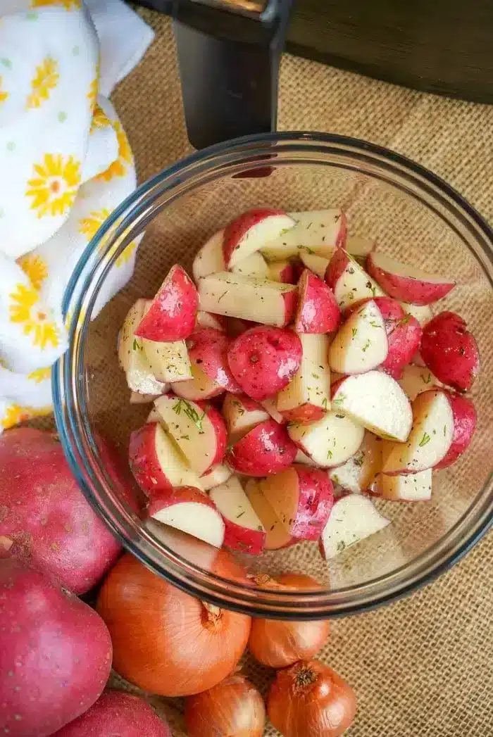 A glass bowl filled with chopped red potatoes seasoned with herbs is placed on a burlap surface, ready to become good air fryer red potatoes. Surrounding the bowl are whole red potatoes, onions, and a white cloth with yellow floral patterns.