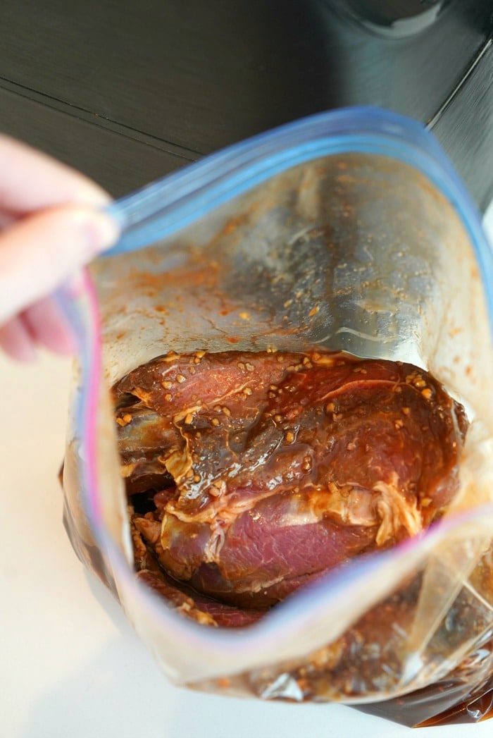 A hand holds an open plastic bag containing raw meat marinated in a red sauce with visible garlic pieces, ready for the air fryer, on a light-colored surface.