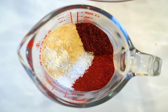 A glass measuring cup viewed from the top, filled with four different spices in partial portions. The spices include a light beige powder, a coarse white powder, and two different shades of red powder, each distinctly separated within the measuring cup—solid for seasoning your next air fryer roast beef.