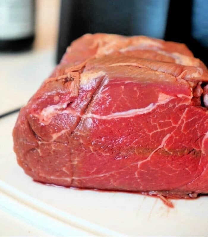 A close-up image of a raw beef roast destined for the air fryer, resting on a white surface. The marbled texture and vibrant red color of the meat are prominently displayed against a dark, blurred background.