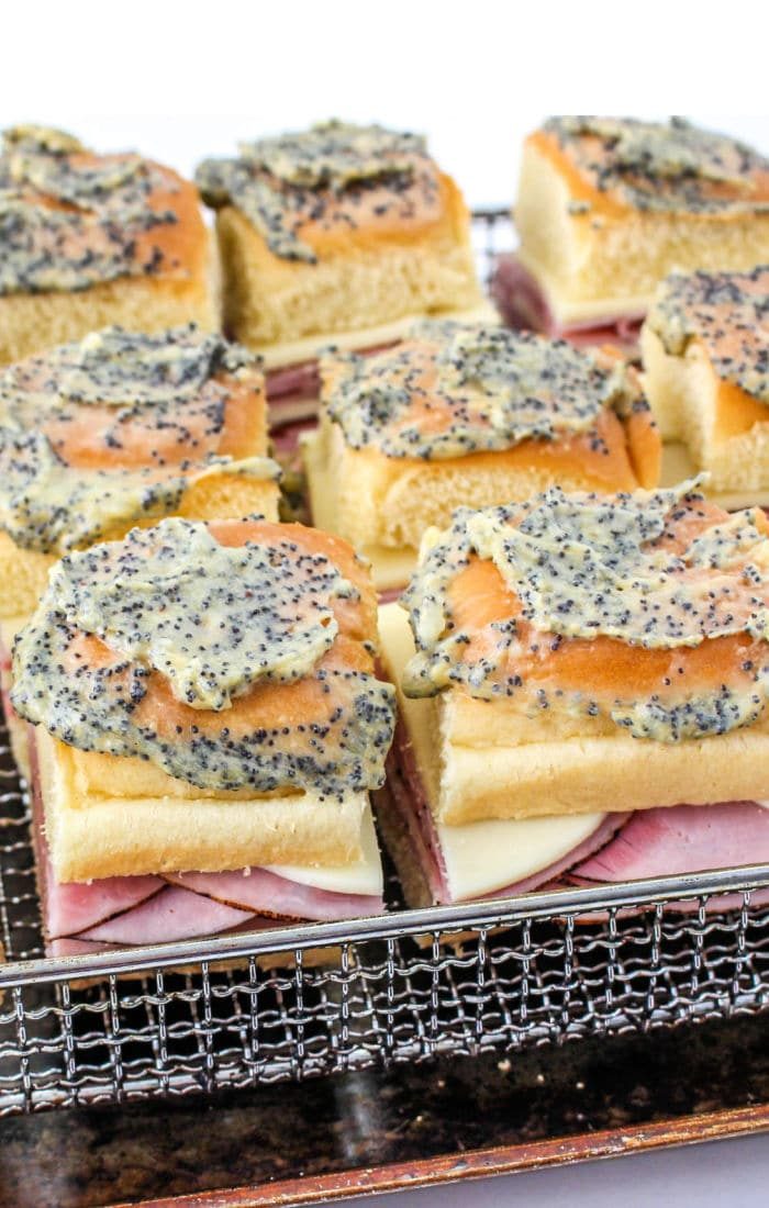 Close-up of ham and cheese sliders on a wire rack, topped with a poppy seed and butter mixture. These air fryer sandwiches boast a layer of melted cheese, perfectly arranged in a grid pattern. The buns are lightly toasted to perfection.