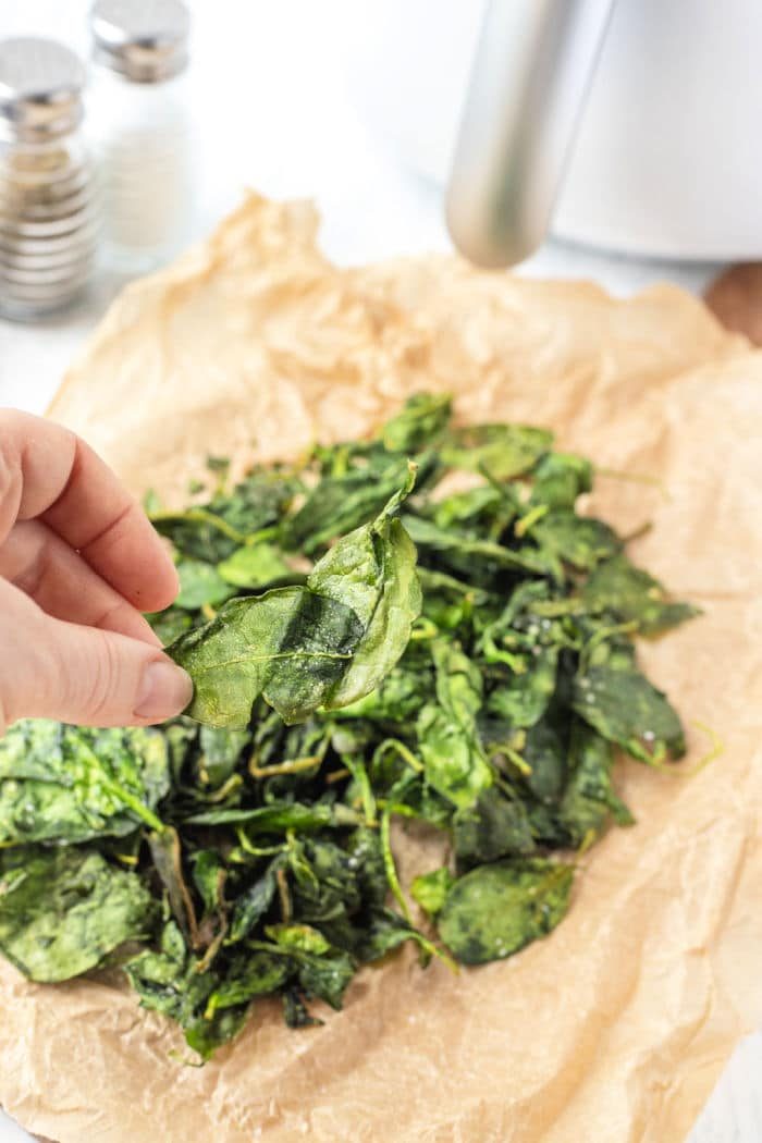 A hand holds a crisp spinach leaf over a pile of perfectly air-fried spinach on parchment paper, showcasing the good result of the air fryer spinach method.