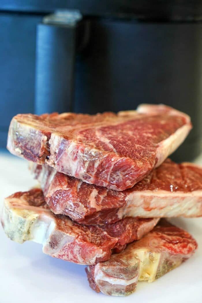 Four frozen raw steaks stacked on a white surface with a black appliance in the background—solid for making juicy air fryer steak bites.