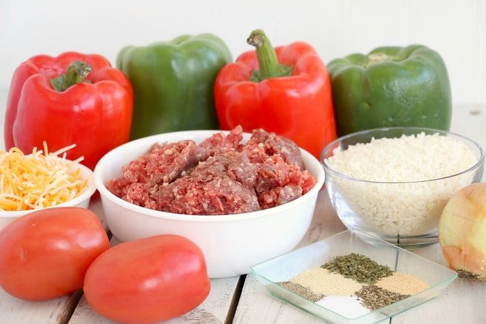 Bell peppers, ground meat, shredded cheese, tomatoes, uncooked rice, spices, and an onion on a white surface—solid ingredients for good air fryer stuffed peppers.