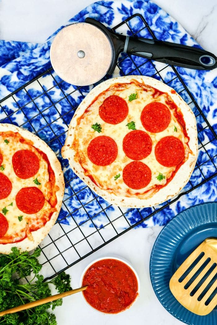 Two Air Fryer Tortilla Pizzas on a cooling rack with a pizza cutter, sauce, and utensils nearby.