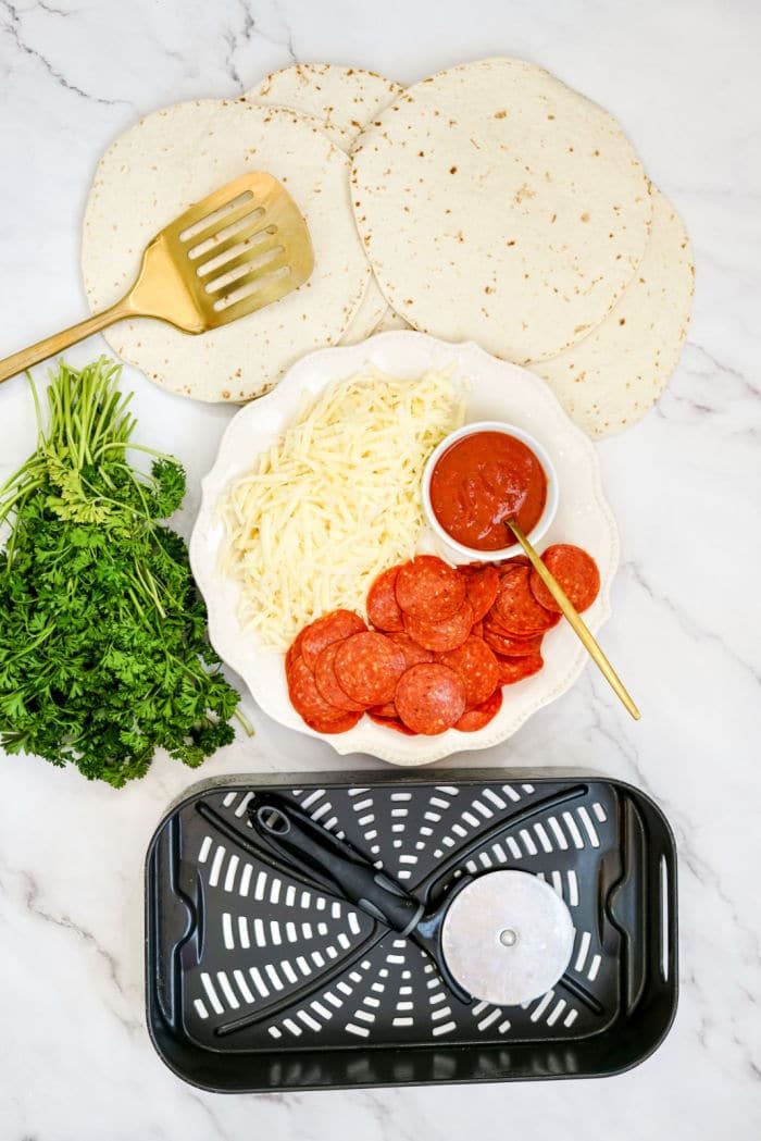 Air Fryer Tortilla Pizza essentials: tortillas, shredded cheese, pepperoni, sauce, parsley, spatula, pizza cutter, and an air fryer basket arranged on the counter.