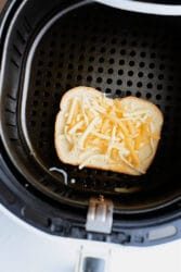 A slice of bread topped with shredded cheese and tuna sits inside the air fryer basket. The air fryer is open, revealing the perforated surface beneath this delectable Air Fryer Tuna Melt.