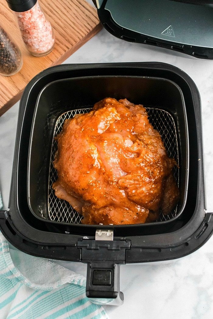 Raw marinated turkey breast placed in an air fryer basket, ready to cook alongside the succulent aroma of air fryer turkey breast. The air fryer lid is open, revealing its contents. A wooden board with salt and pepper shakers is visible in the background, with a blue and white cloth partially seen at the bottom.