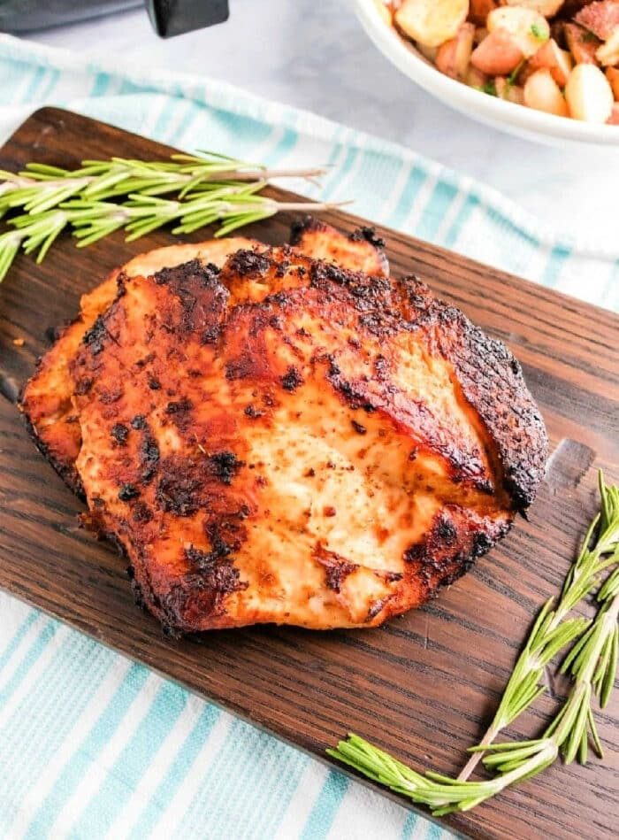 air fryer boneless turkey breast with a crispy exterior rests on a wooden board, adorned with sprigs of rosemary. In the background, an air fryer turkey breast and a bowl of roasted potatoes sit charmingly on a striped cloth.