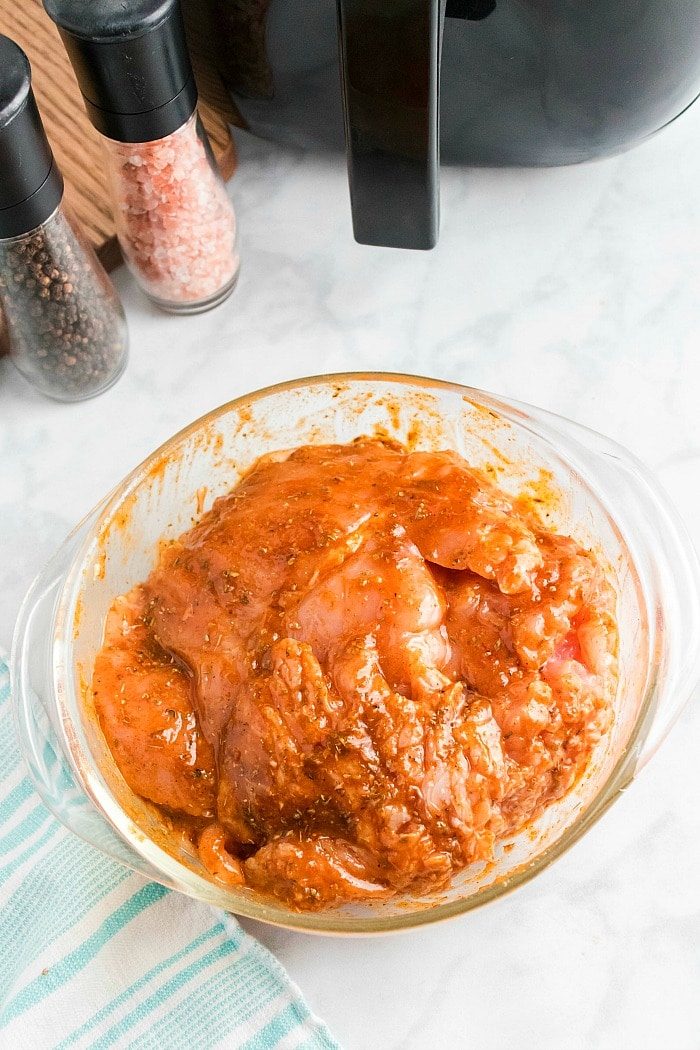A glass bowl filled with raw boneless turkey breast marinating in a reddish sauce is placed on a marble countertop. Nearby, an air fryer hints at future meals, alongside salt and pepper grinders, a striped cloth, and a black appliance.