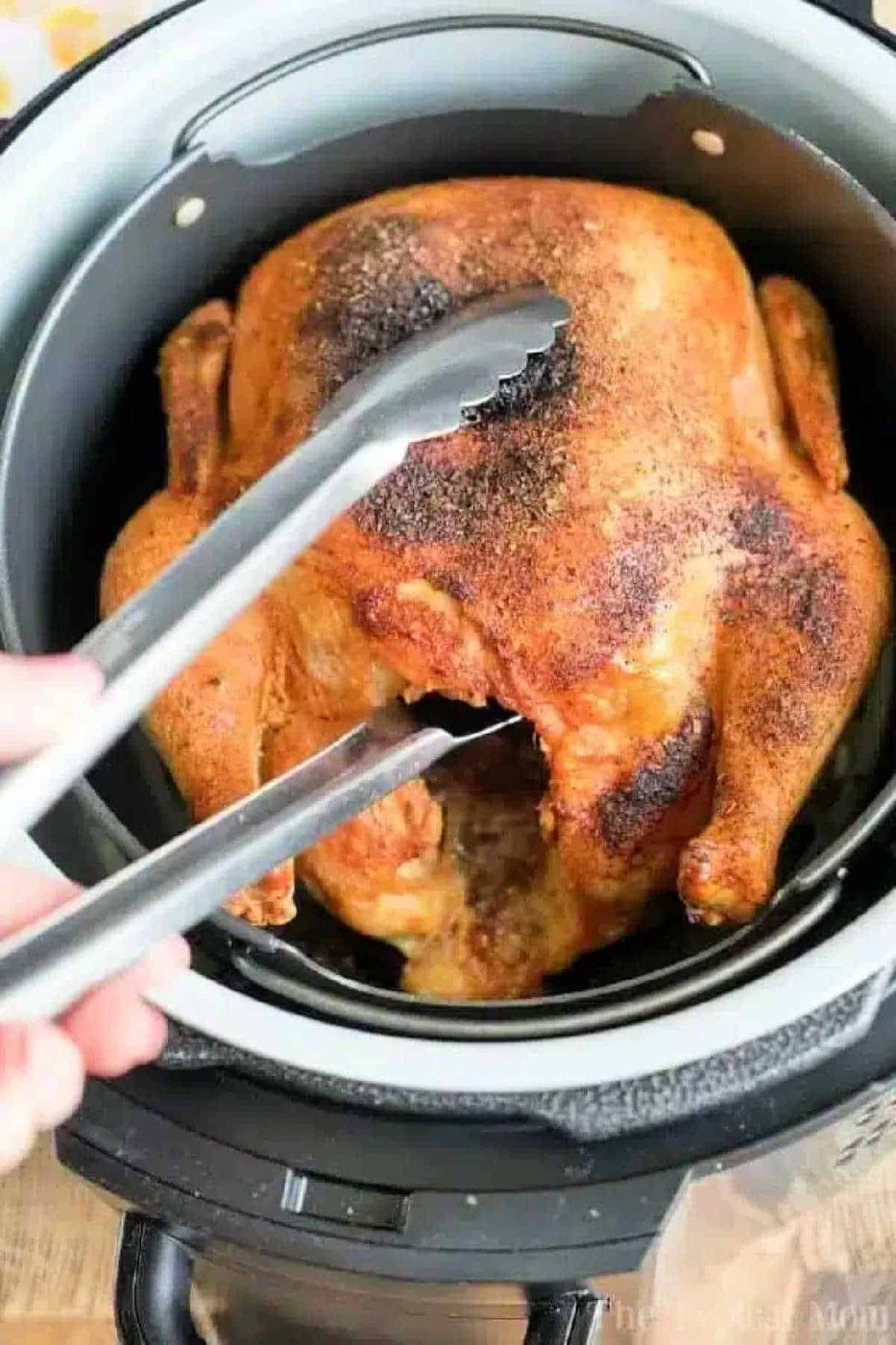 An air fryer whole chicken sits in the basket, perfectly cooked and golden, as metal tongs lift it up for serving.