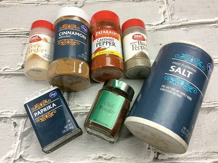 A collection of spice containers on a white brick surface, solid for an air fryer wings recipe, includes garlic powder, ground cinnamon, cayenne pepper, black pepper, paprika, chili powder, and salt. Each container showcases distinct branding and labels.