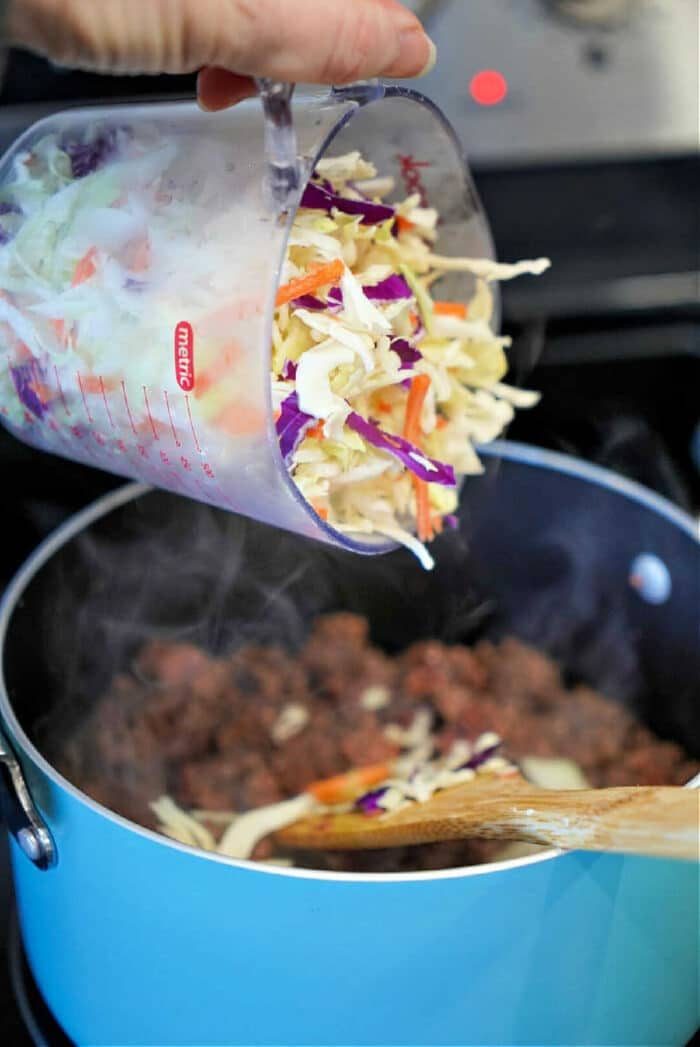 Hand pouring shredded cabbage mix from a measuring cup into a blue pot with cooked ground meat, solid for a savory filling in homemade air fryer won tons.