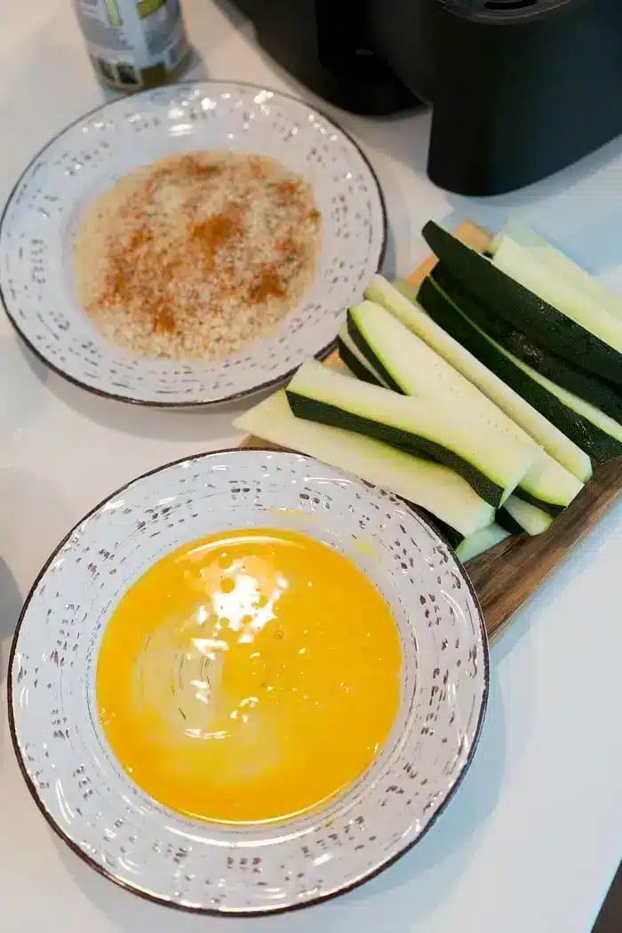 Two bowls on a white surface, one with beaten eggs, the other with a breadcrumb mixture. Next to the bowls are sliced zucchini on a cutting board, ready to become good air fryer zucchini fries.