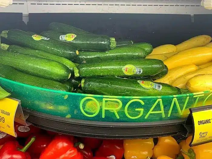 A grocery store display features green zucchini with produce stickers and yellow squash, solid for making air fryer zucchini fries. A partially visible yellow sign reads u0022ORGANIC.u0022 Red bell peppers are visible in the bottom of the frame.