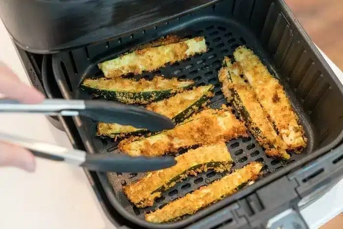 A person uses tongs to remove crispy air fryer zucchini fries from the basket. The zucchini sticks are coated in a golden-brown breading and evenly spaced in the black basket, with the air fryer lid open to reveal the good contents.