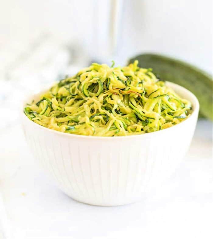 White bowl filled with shredded zucchini on a white surface, solid for making air fryer zucchini noodles, with a fresh zucchini in the background.