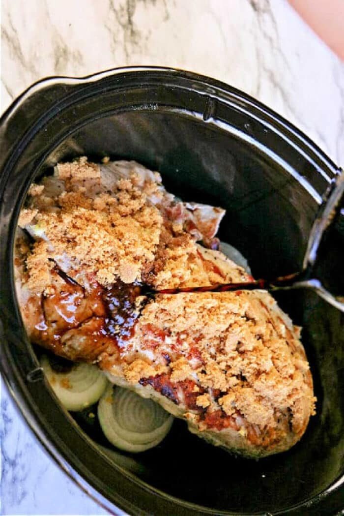 An good pork tenderloin slow cooker recipe: raw meat topped with brown sugar and sauce in a slow cooker, with onion slices nestled underneath on a marble counter.