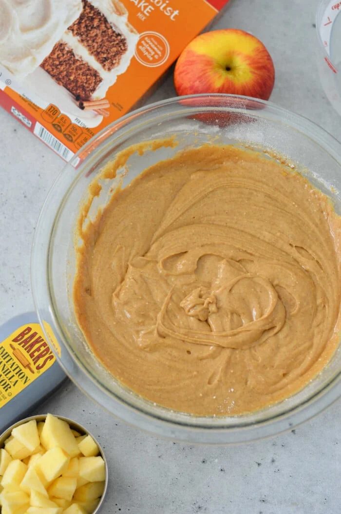 A glass bowl filled with cake batter sits gracefully on the countertop, surrounded by a box of cake mix, chopped apples, a whole apple, and a measuring cup—solid ingredients for an effortless apple cake.