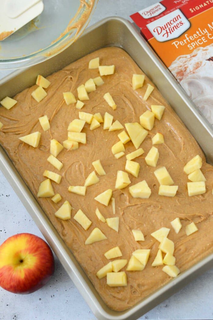 A baking pan filled with apple cake batter, nestled between a fresh apple and a convenient cake mix box, is ready to transform into a good apple cake with cake mix.