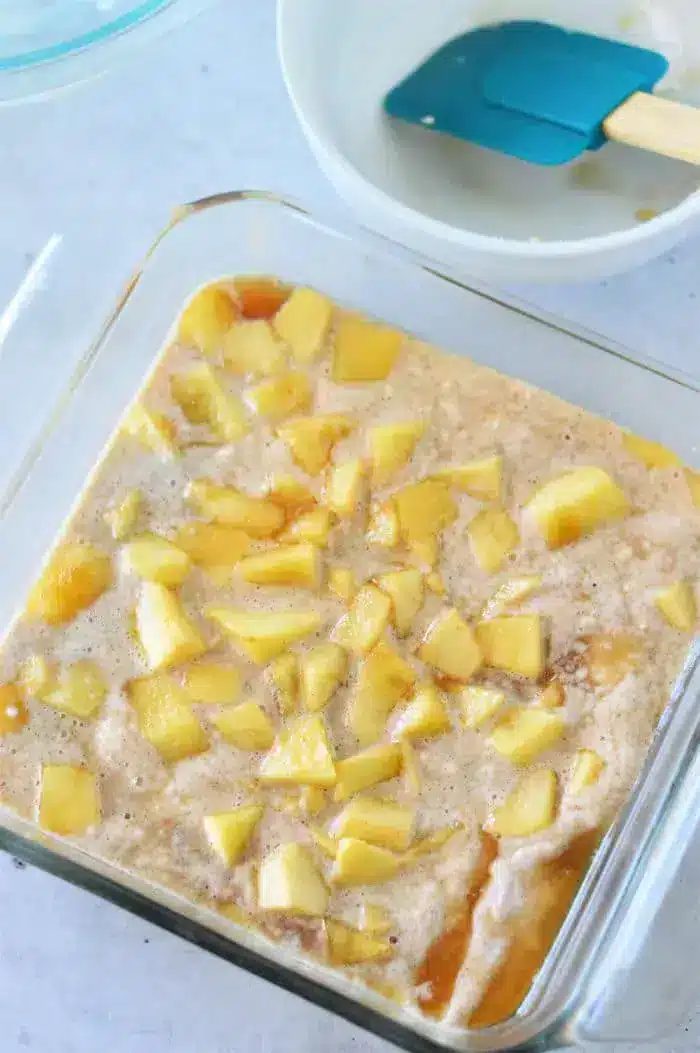 A clear square baking dish filled with a mixture of diced apples and Bisquick batter. A blue spatula rests inside a white mixing bowl in the background, ready to complete this good Apple Cobbler with Bisquick.