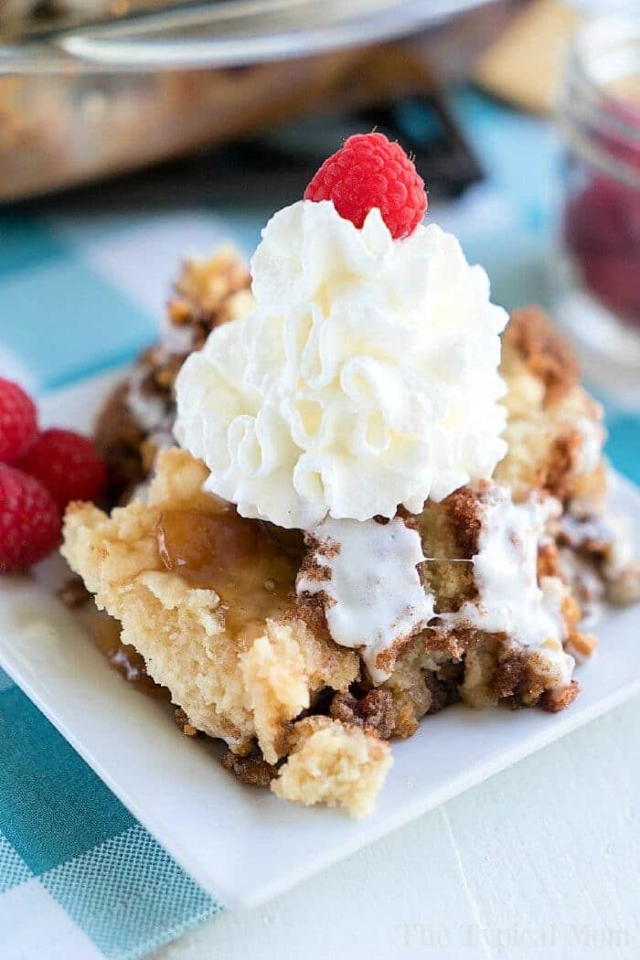 A slice of apple spice dump cake topped with whipped cream and a raspberry on a white plate, with more raspberries on the side.