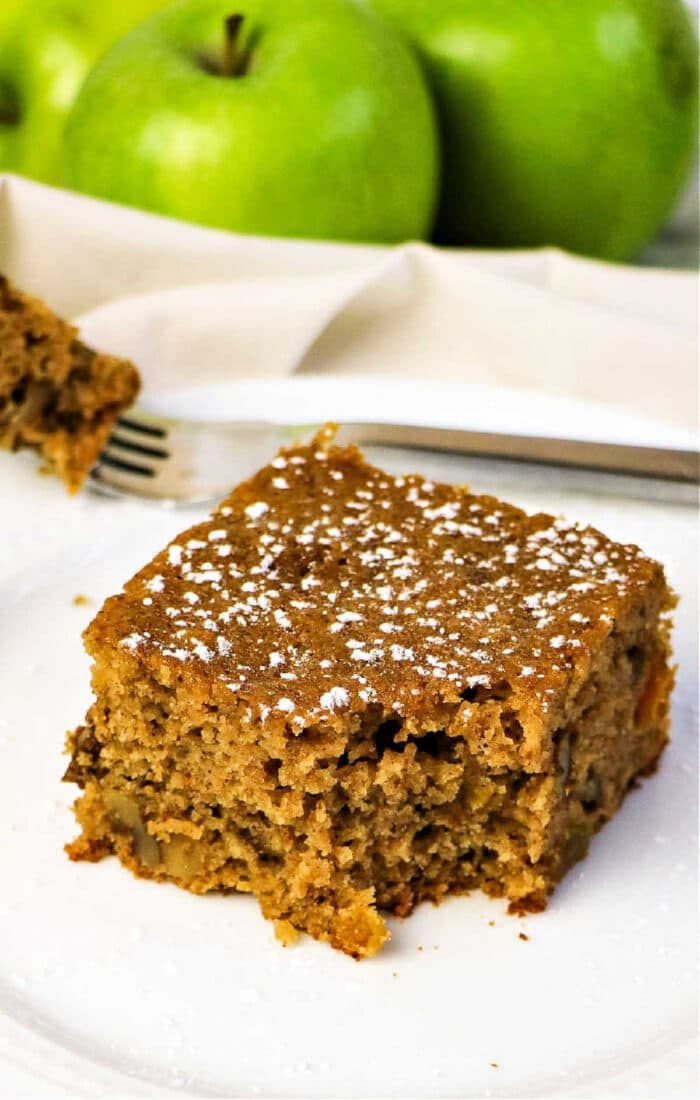 A slice of applesauce spice cake dusted with powdered sugar sits on a white plate, with green apples in the background.