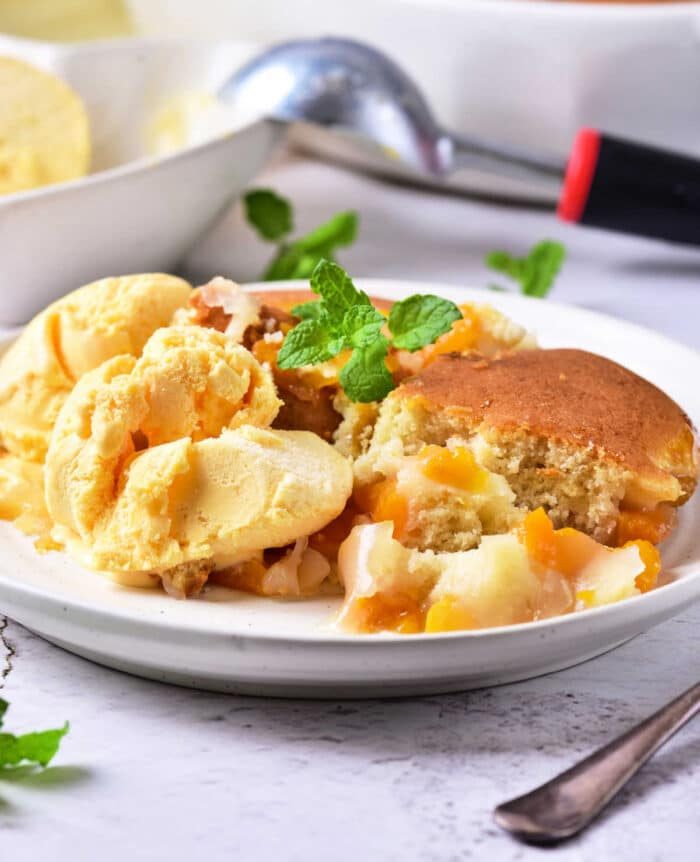 A plate of apricot cobbler with vanilla ice cream, garnished with mint leaves, sits invitingly next to a serving spoon.