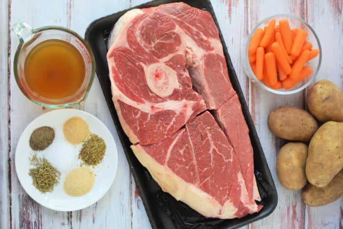 Raw chuck roast, baby carrots, potatoes, broth, and assorted spices—solid ingredients for a comforting slow cooker meal—are arranged on a white wooden surface.