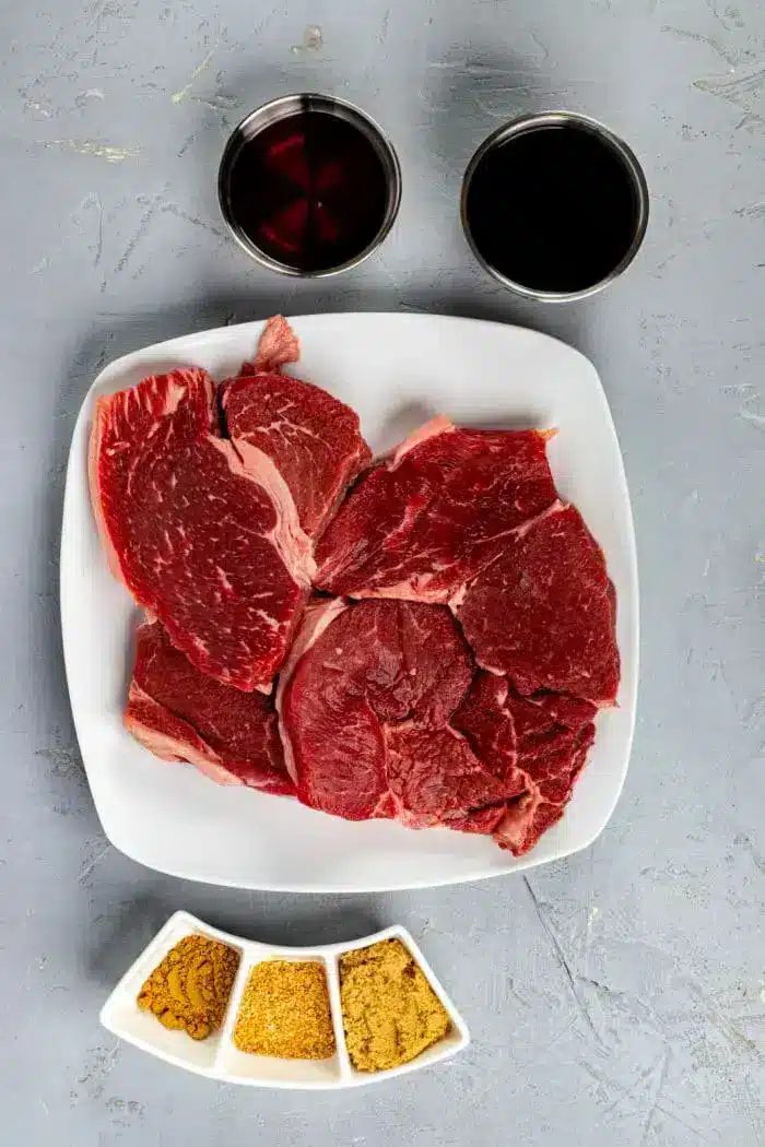 A square white plate with several pieces of raw red beef sits on a textured gray surface, suggesting a solid base for an Asian steak marinade. Above the plate are two cups, one filled with a dark liquid and the other with a reddish liquid. Below the plate are three small white dishes with various spices.
