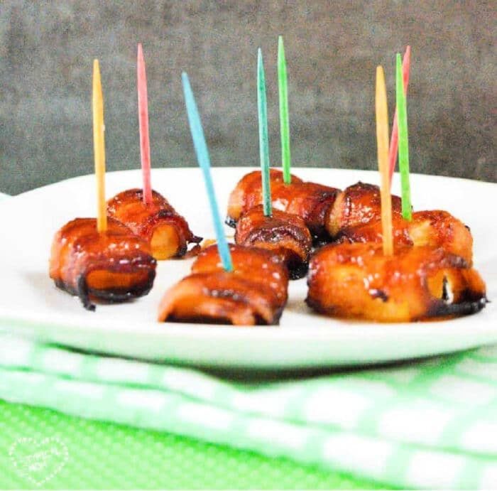 Bacon-wrapped pineapple bites with colorful toothpicks adorn a white plate, elegantly set on a green-checkered cloth.
