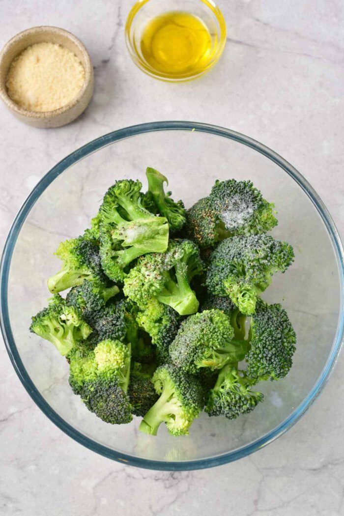 A glass bowl of raw broccoli florets with seasonings, solid to bake frozen broccoli, sits next to bowls of olive oil and grated cheese.