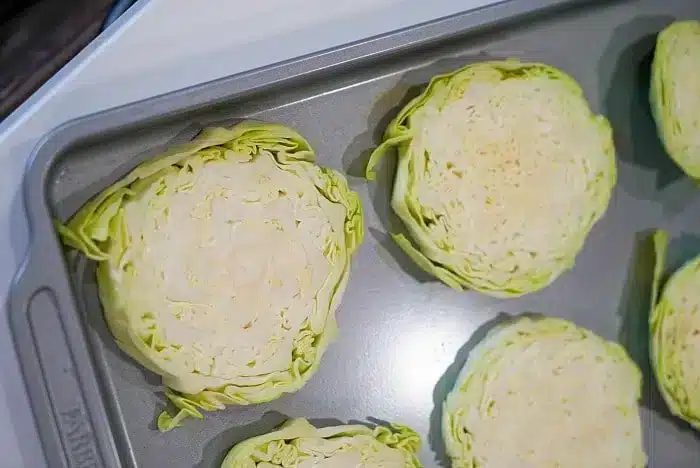 Slices of cabbage arranged on a baking sheet, ready for roasting or baking into good baked cabbage steaks. The cabbage is cut into thick rounds, showing the layers of leaves within each slice. The baking sheet is placed on a countertop.
