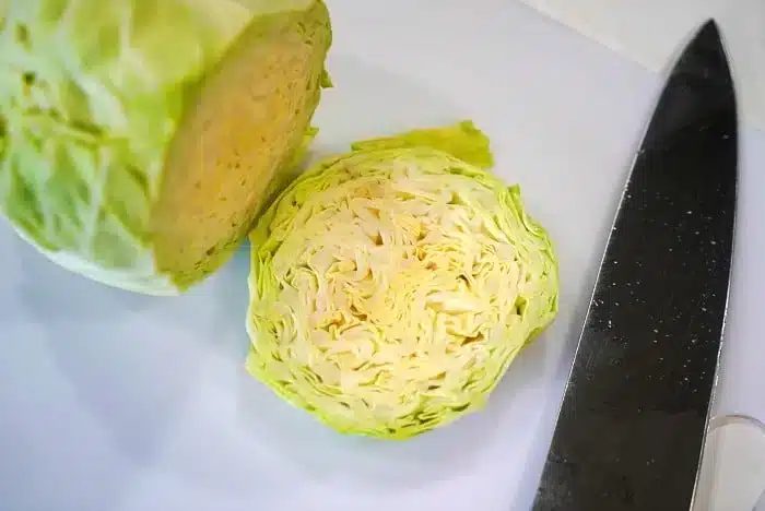 A halved cabbage is placed on a white surface next to a large kitchen knife, ready to be transformed into good baked cabbage steaks. The inner layers of the cabbage are visible, showcasing its tightly packed, light green and white leaves.