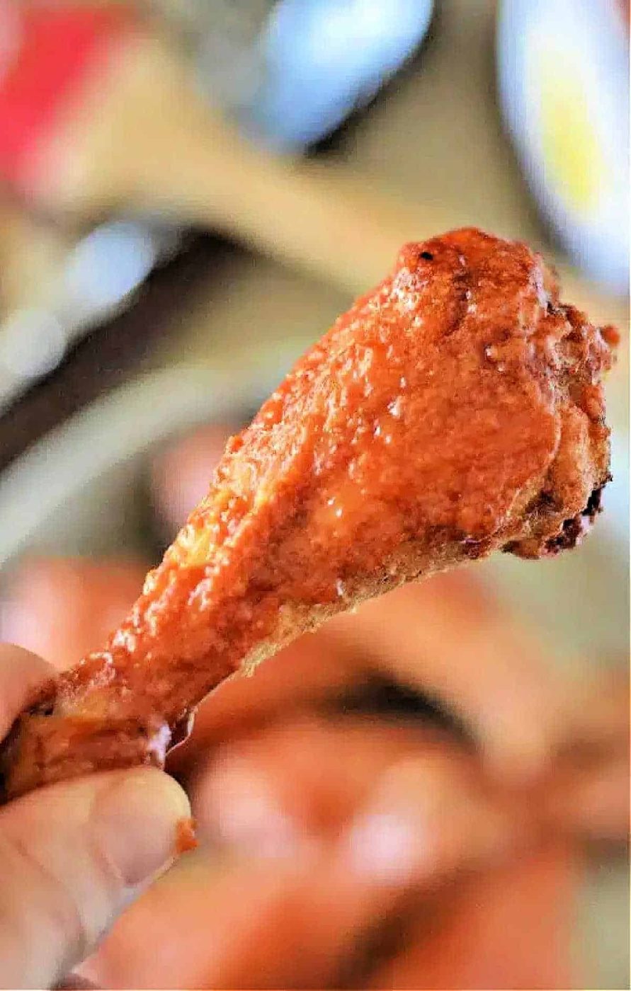 A hand holding a crispy, saucy baked chicken leg, reminiscent of buffalo chicken drumsticks, with a blurred background.