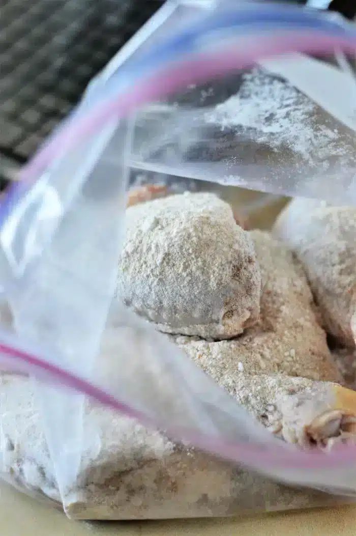 An open plastic bag containing raw, floured chicken pieces rests on a kitchen counter. The chicken pieces are coated with flour, ready for further preparation like turning them into good baked chicken legs. The background shows a partial view of a dark kitchen appliance.