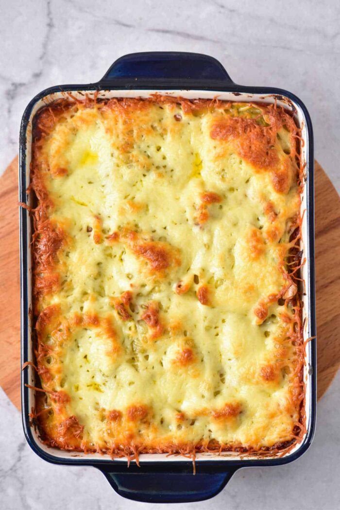 Baked cheesy pasta casserole made with baked frozen ravioli, featuring a golden, bubbly top in a rectangular blue baking dish on a wooden board.