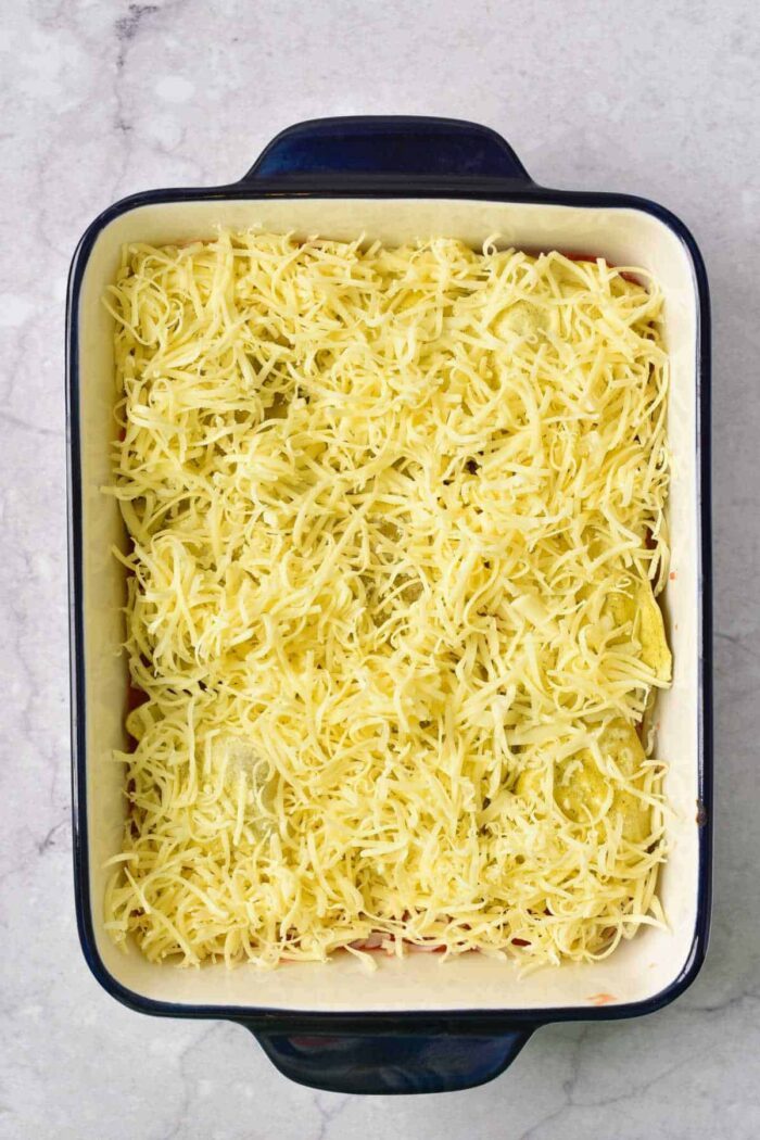 Rectangular baking dish filled with shredded cheese atop a layered casserole, featuring Baked Frozen Ravioli, ready to be baked.