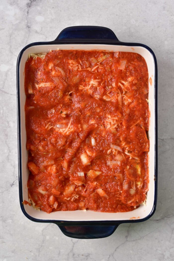 Rectangular baking dish filled with baked frozen ravioli, topped with shredded cheese and tomato sauce mixture, on a gray marble surface.