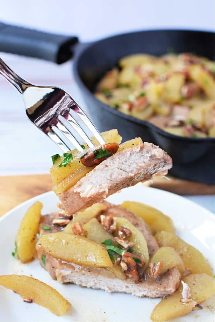 A fork holds a bite of baked pork chop topped with cooked apples and pecans above a plated serving and a skillet, solid for showcasing this comforting Baked Pork Chops with Apples recipe.